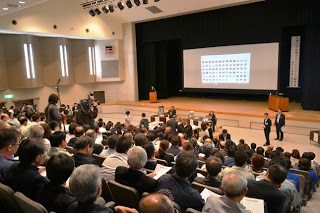 明るい照明の下、大勢の人々が座ってステージを見つめる大きな講堂で、ステージには演台とに映像が映し出された大きなスクリーンがあり、数名がステージ前方に立ってイベントの準備や参加をしている様子の写真