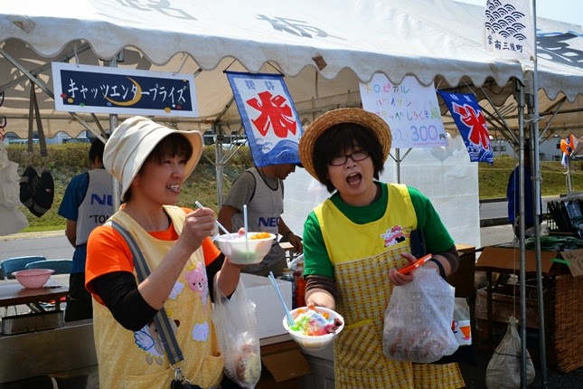 「キャッツエンタープライズ」と書かれた販売ブースのテントの上に、手書きで「かき氷 300円」と書かれた看板が掲げられ、屋台の前でエプロンを着用し、商品を購入した袋をぶら下げた二人の女性が、色とりどりのかき氷を手に持って立っている写真