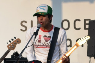 マイクの前で緑と白の帽子をかぶり、歌いながらエレキギターを弾いているよっちゃんの写真