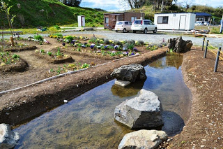 整備された小さな池の横に花壇や2台の自動車、プレハブの建物が見える写真