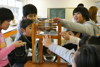 子どもたちが教室の中で、木製の糸取り機を囲みながら、絹の糸取り体験をして楽しそうに触れている様子が写っている写真