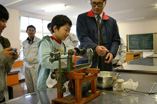 子どもが絹糸を取る機械を自分で操作し、隣で男性が指導している様子の写真