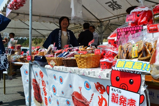 テントの下にある祭りの屋台で、タコのキャラクターがデザインされたイラストと「ゆめ多幸」と書かれたクロスの上に、色とりどりのパッケージや「新商品!!」と書かれた看板が飾られ、せんべいなどの包装された食品がかごに並び、法被姿の女性スタッフが笑顔で写っている写真