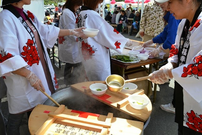 法被を着た女性たちが、白い容器に大鍋からお吸い物をよそい、来場者に振る舞っている様子の写真
