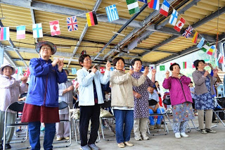 国旗が飾られた屋根の下で高齢の女性たちが椅子から立ち上がり、人差し指を上げた両手を左側に向けて踊りに参加している様子の写真