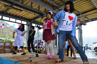 屋根付きのステージの上で2人の女性が踊るなか、中央のメイド服に仮装した女性が歌を歌っている様子の写真