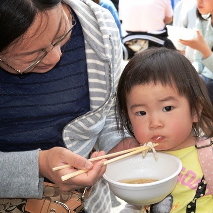 母親が小さな子どもに白い器から小さな魚を箸で食べさせていて、子どもがカメラを見ながら食べている写真