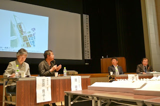 講堂のステージ上で、テーブルに座った4人のパネリストが建築図面や地図を映した大きなスクリーンを背に、討論を行っている様子を写した写真