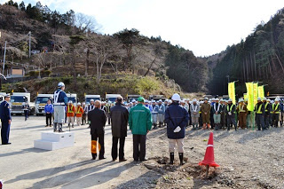 山や木々が広がる背景には車両が並び、右側には黄色い旗を持ち、ヘルメットと安全ベストを着た人々が屋外に並び、台の上の人物が挨拶を行っている様子の写真
