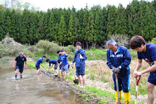 川のように水のたまった田んぼの縁に、大人や若者たちがはだしや長靴を履いてスコップなどの道具を使いながら整地作業をしている農作業中の写真