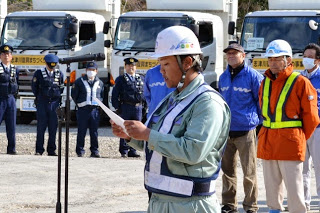 複数のトラックが並んだ前に、制服や作業着を着た人々が屋外に集まり、マイクスタンドの前に立つ男性が紙を持って話している様子の写真