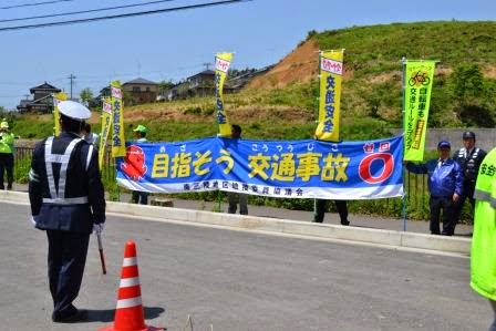 黄色ののぼり旗や「目指そう交通事故ゼロ」と書かれた青い横断幕を数人が掲げており、警察官も配置されている交通安全啓発活動の様子の写真