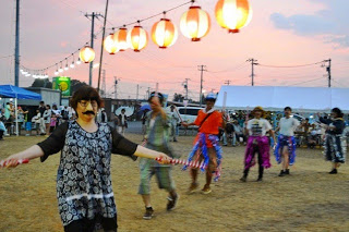 夕暮れの会場で提灯の下、派手な衣装の人たちが踊りながら盆踊りを楽しんでいる写真