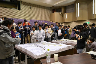 屋内の講堂で、大きなテーブルを囲んで人々が建築や工学の図面と思われる資料を熱心に見ている様子を写した写真