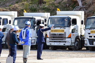 白いトラックの前にヘルメットを被った人々が集まり、制服姿の警官が右手を真っすぐ前に伸ばし指示を出している様子が見られる写真