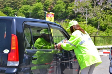 黄色い安全ジャンパーを着た女性が黒い車の運転手に交通安全啓発のチラシなどを手渡している活動中の写真