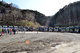 澄んだ空が広がり緑豊かな林を背景に、砂利の広場に半円状に並んだ建設用トラックと、その左側に人々が並んで立っている写真