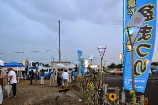 夏祭りの入り口付近で、青い旗やヒマワリが並ぶ通りを多くの人が傘を差して歩いている写真