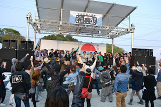 野外のステージで多くの人々が手を挙げて盛り上がっている様子を、観客の背中越しに撮影した写真