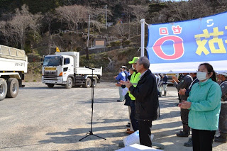 前方にはマイクスタンド、後方には大きな横断幕が掲げられた屋外で、出発する2台のトラックに向けて、人々が拍手をしている様子が写された写真