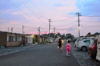 夕焼けの空の下、仮設住宅の並ぶ通りを浴衣姿の子どもと大人が歩いている後ろ姿の写真
