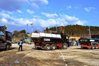 空には雲が広がり、木々が並んだ背景に、複数のダンプトラックと作業員が土の地面に白い印を付けた広場から出発している様子の写真