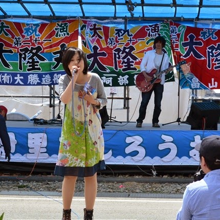 大漁旗が飾られている野外ステージで、女性がマイクを持って歌い、後ろで男性がギターを弾いているライブ中の写真