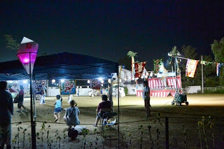 夜の盆踊り会場で、提灯や紅白幕に照らされた櫓を中心に、見物客がテントの下や芝生に座っている様子の写真