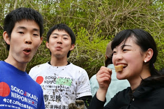 若い男性二人と女性一人が屋外で楽しそうに食べ物を頬張っていて、背景には緑の木々が生い茂っている自然の中での休憩時間の写真