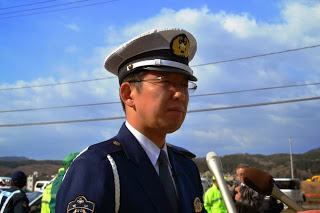 青空の下に立ち、マイクを向けられている白い帽子をかぶった制服姿の警察官をアップで撮影した写真