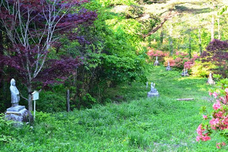 ツツジや木々に囲まれた緑の草に覆われたなだらかな斜面に、様々な形をした複数の観音像が点在する写真