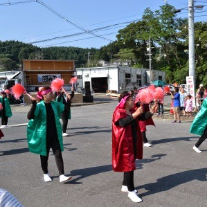 両手にピンクのポンポンを手に持った緑と赤の光沢ある衣装を着たグループが、観客の前でにこやかに踊っているトコヤッサイコンテストの様子の写真