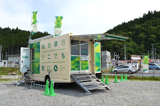 「仙台銀行 どこでも窓口」と書かれた側面を開いて、移動型銀行車が仮設の窓口を設置している様子を左側から写した写真