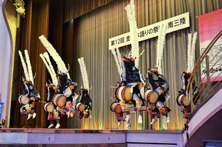 横断幕が掲げられたステージの下で、太鼓を叩きながら踊る複数の演者が一斉に高く跳び上がっている写真