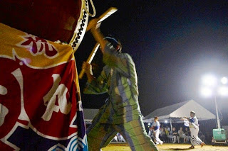 夜の盆踊り会場で、照明の下で法被を着た男性が大きな和太鼓を力強く叩いている瞬間をとらえた写真