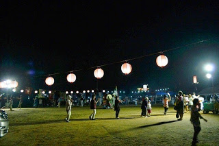 提灯の明かりが吊るされた夜の広場で、人々がゆったりと踊りながら盆踊りを楽しんでいる様子の写真