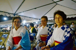 夜の会場のテントの下で、赤い帯を締めた色違いの浴衣を着た女性たちが並んで座り笑顔を見せている写真