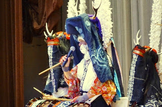 鹿の角のような飾りと色鮮やかな衣装を身につけた人物がマイクを持って話している写真