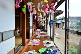 古民家の縁側にカラフルな布や紙で作られた作品が天井から吊るされ、床にも作品が並べられている写真