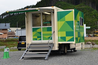 緑の幾何学模様が描かれた移動銀行車を後方左側から見て、後部入口に階段が設置されている写真