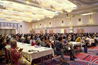 ホテルの大広間で、多数の参加者がテーブル席に座り、舞台に注目して一斉に拍手を送っている写真