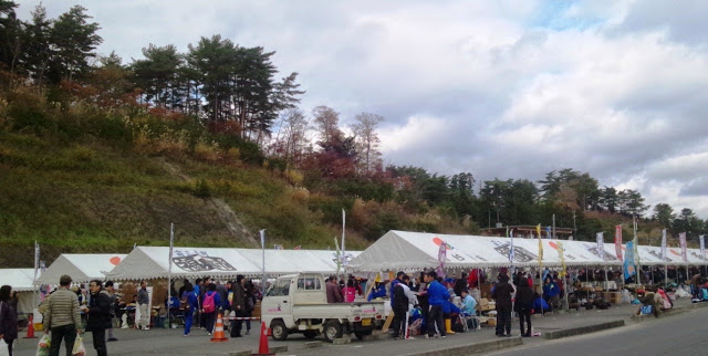 小高い山の麓に並んだ白いテントで祭りが開かれ、大勢の人が集まってにぎわっている写真
