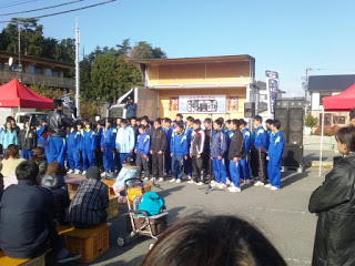 屋外ステージの前で青いジャージ姿の生徒たちが整列している写真