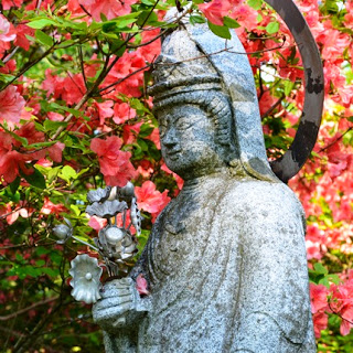 赤いツツジが咲き誇る中で、冠をかぶった観音像が花の飾りを手に持ち、やさしい表情で立っている写真