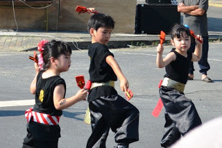 黒い衣装に金色の帯をつけた小さな子どもたちが、手に鳴子を持って元気に踊っている写真
