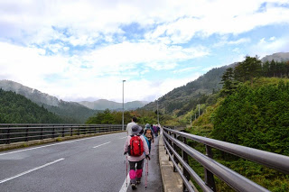 山々が続く風景の中、舗装された橋の上を赤いリュックを背負った女性を最後に、数人の参加者が列をなして歩いている様子を後方から撮影した写真