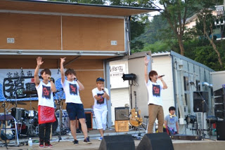 後ろにはバンド機材が置かれている仮設ステージの上で、4人の男性グループがマイクを持って手を挙げながら歌っている写真