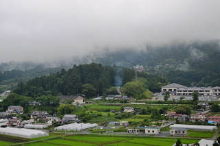 山のふもとに広がる農村集落とビニールハウス、そして霧がかかった森林が広がる風景写真