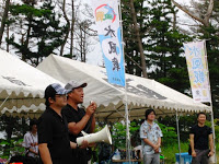 「水風戦」と書かれた旗が掲げられたテントの前に立った男性のうち一人が拡声器を使って話をしている写真