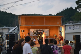 バンドの演奏に合わせて女性ボーカルがステージの中央に立ち歌っており、夕暮れの中で観客が音楽に聴き入っている音楽ステージの写真